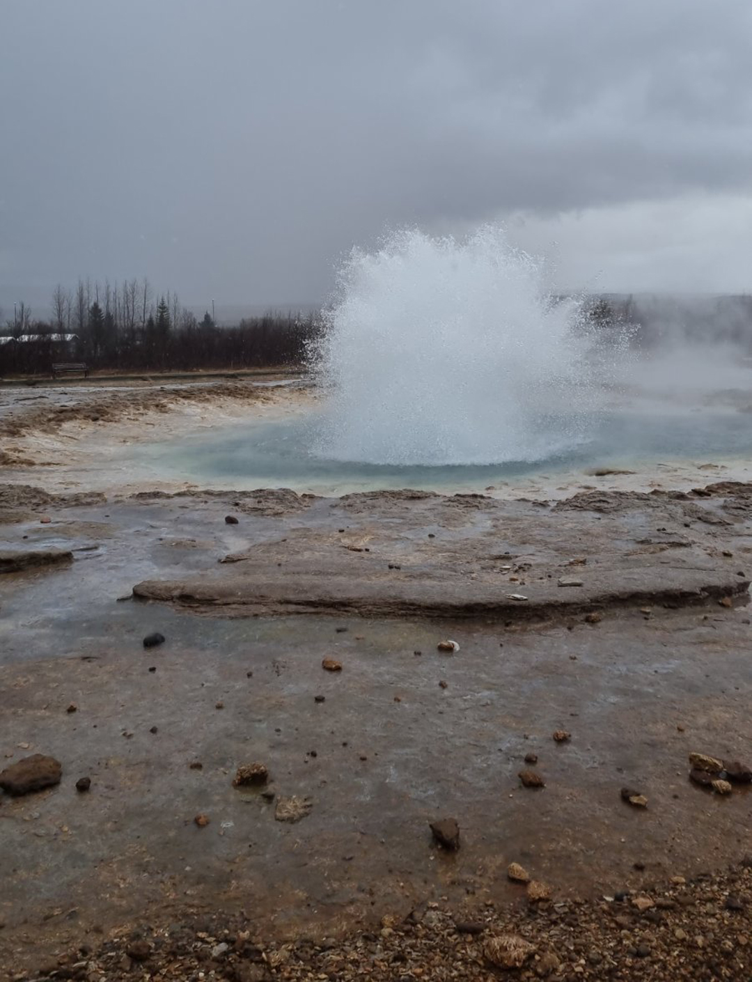Gejzir Strokkur v trenutku močnega izbruha, ko vroča voda brizga visoko v zrak nad geotermalnim poljem.
