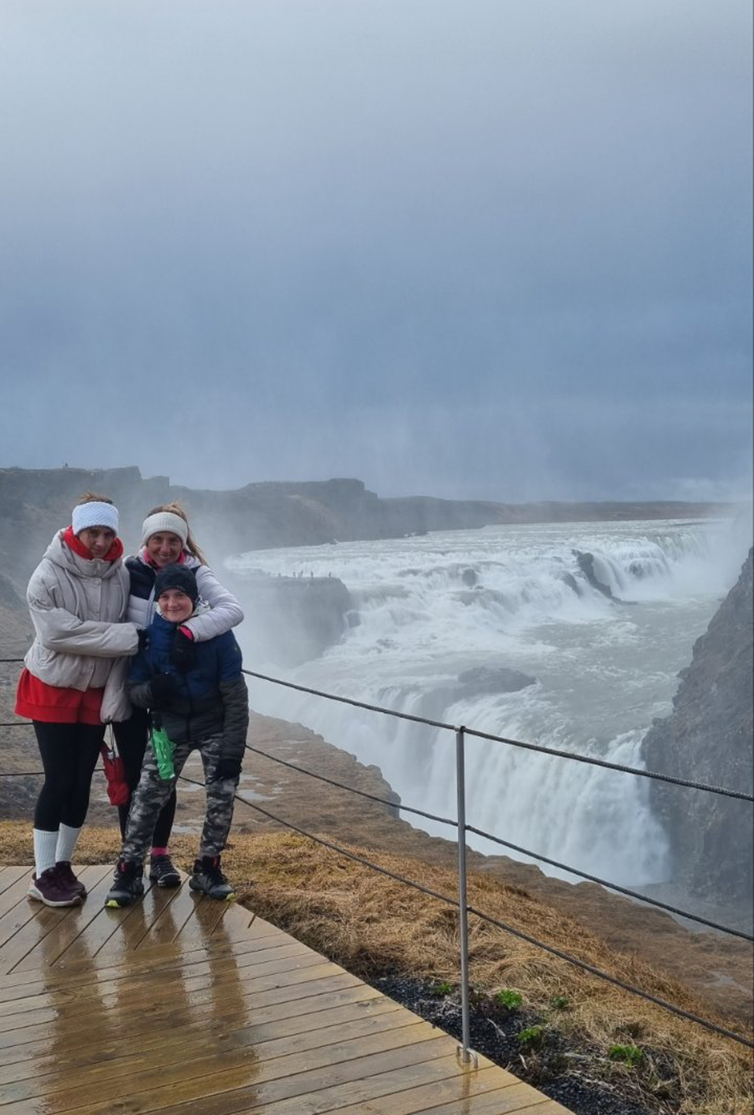Družina stoji ob razgledni točki nad mogočnim slapom Gullfoss, obdanem z vetrom, meglico in dramatično pokrajino.
